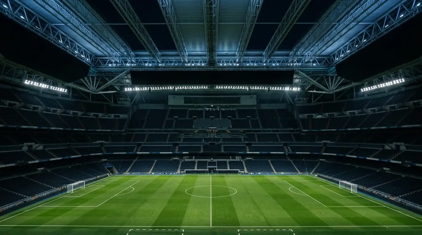 An editorial wide shot of a grand modern football stadium interior with a closed retractable roof, empty pitch immaculately lit, tiered seating rising into shadow, a sense of anticipation before a major match